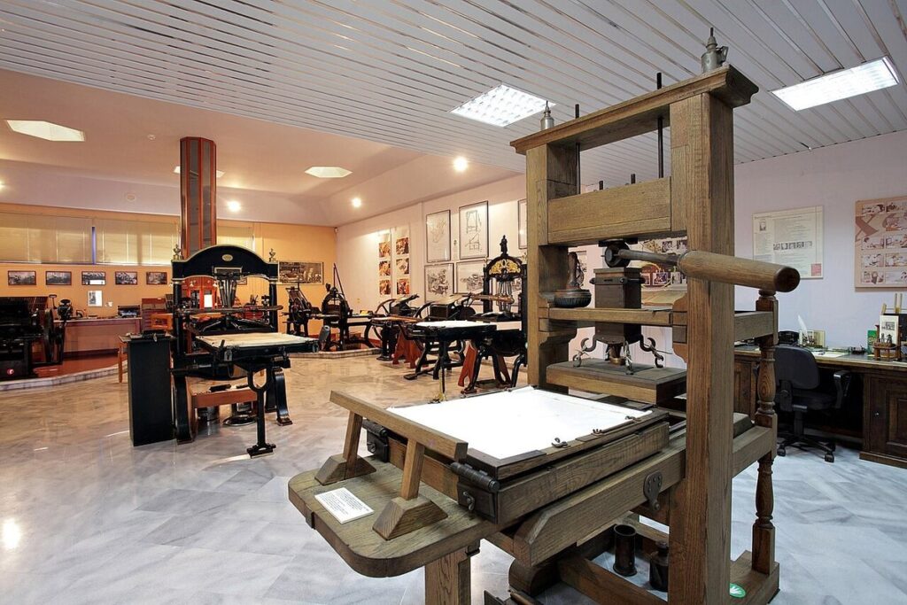 Replica of a Gutenberg-era wooden printing press in front of 19th-century cast-iron presses at a museum of typography