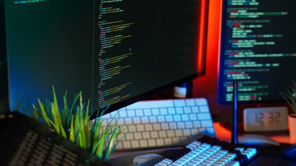 A developer's desk at night with source code on two monitors, a keyboard and a small plant