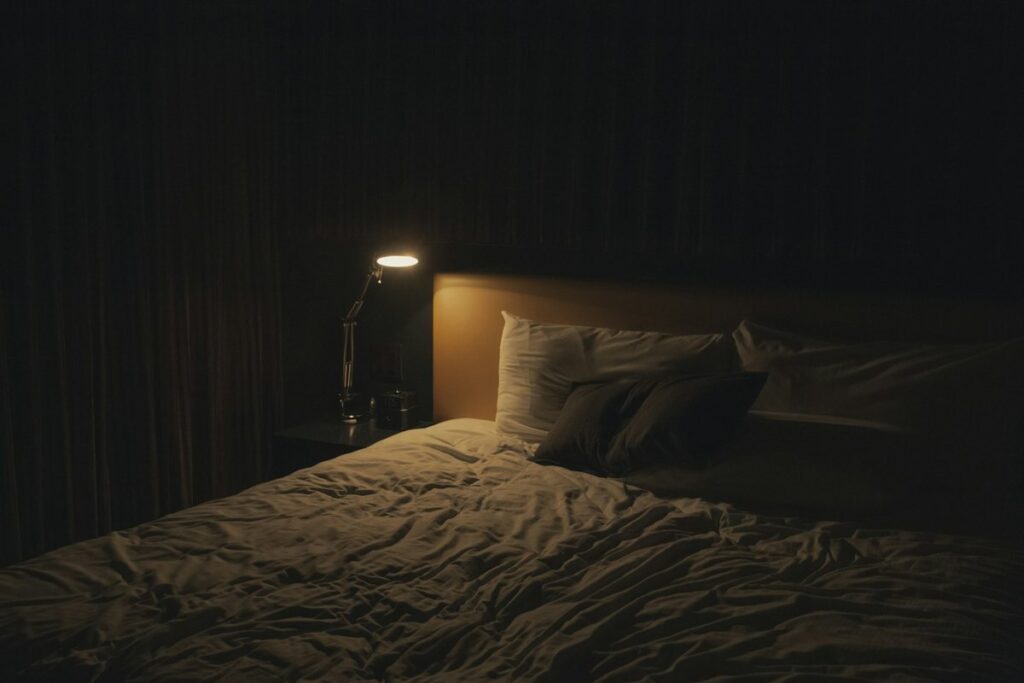 A dim bedroom at night, with a bedside lamp glowing over an unmade bed