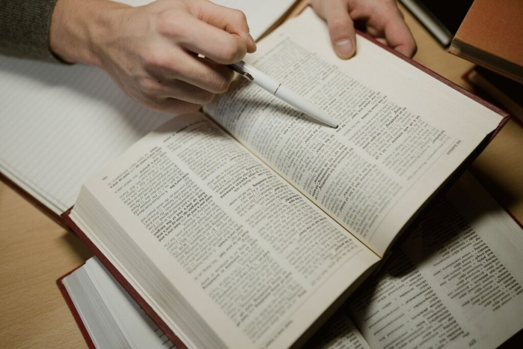 Hands holding a pen over an open reference book, with several other books open on the desk
