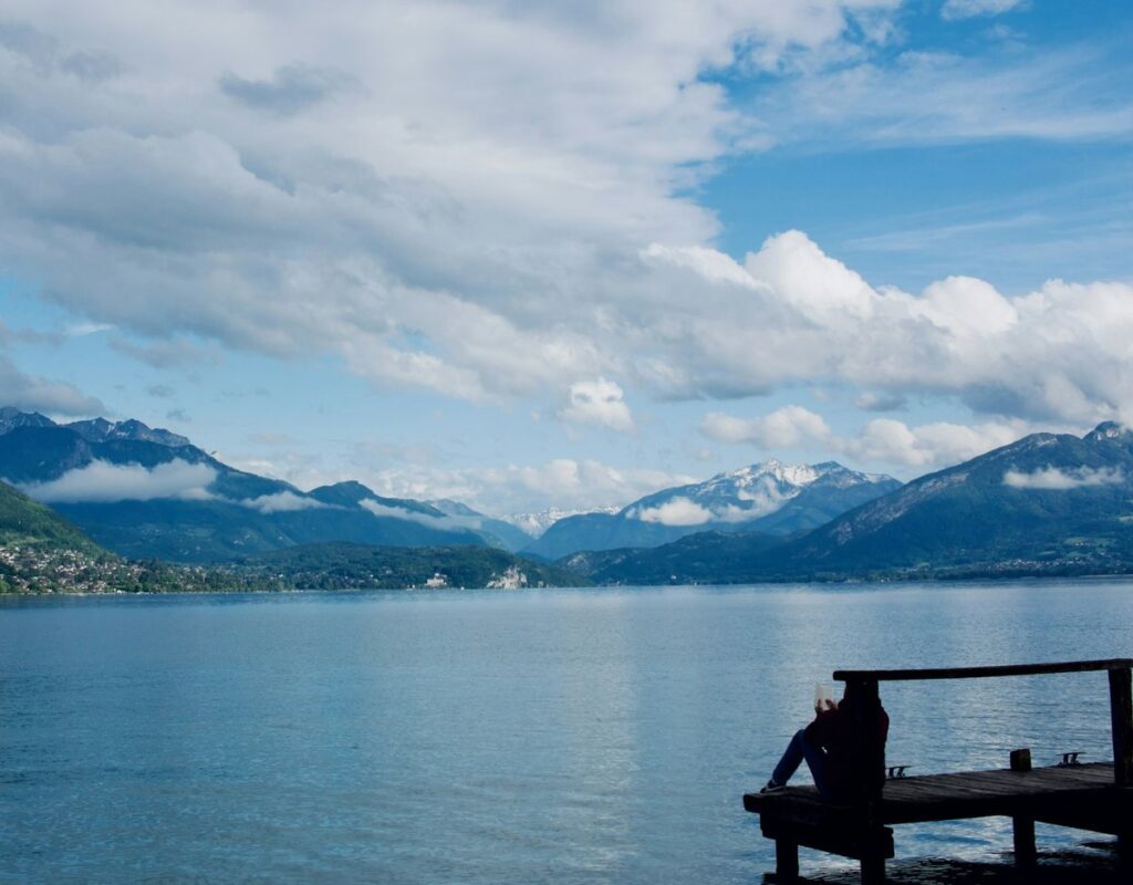 A lone figure sits reading on a wooden jetty beside a calm mountain lake under a bright, cloud-streaked sky