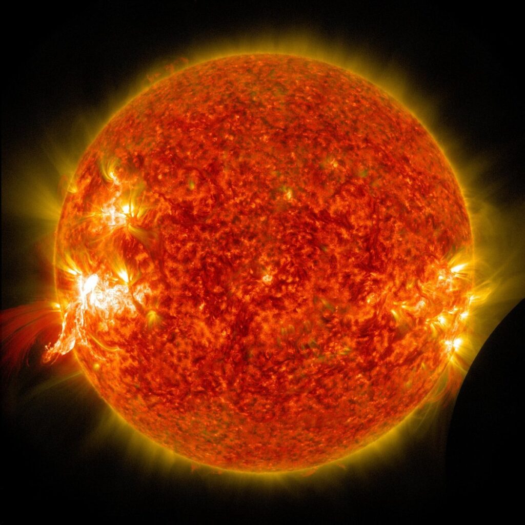 The Sun glowing orange as a solar flare erupts on its left edge, seen by NASA's Solar Dynamics Observatory with the Moon's dark edge at lower right