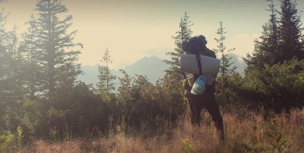 A lone backpacker with a heavy pack and sleeping mat looks out over forested wilderness toward distant mountains