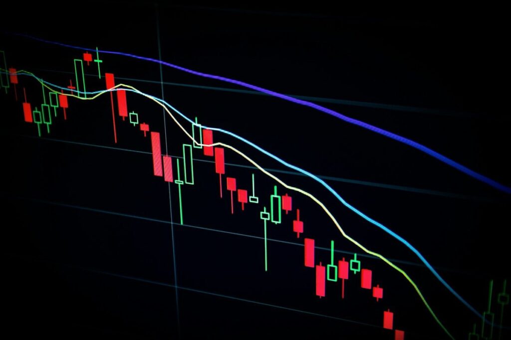 Red and green candlestick stock chart with moving-average lines trending down on a dark screen
