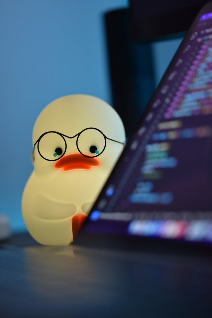 A duck figure wearing glasses beside a laptop showing code, a nod to rubber duck debugging