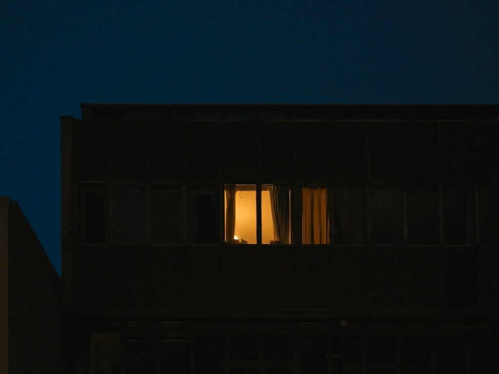 A single warmly lit, curtained window in an otherwise dark apartment building at night