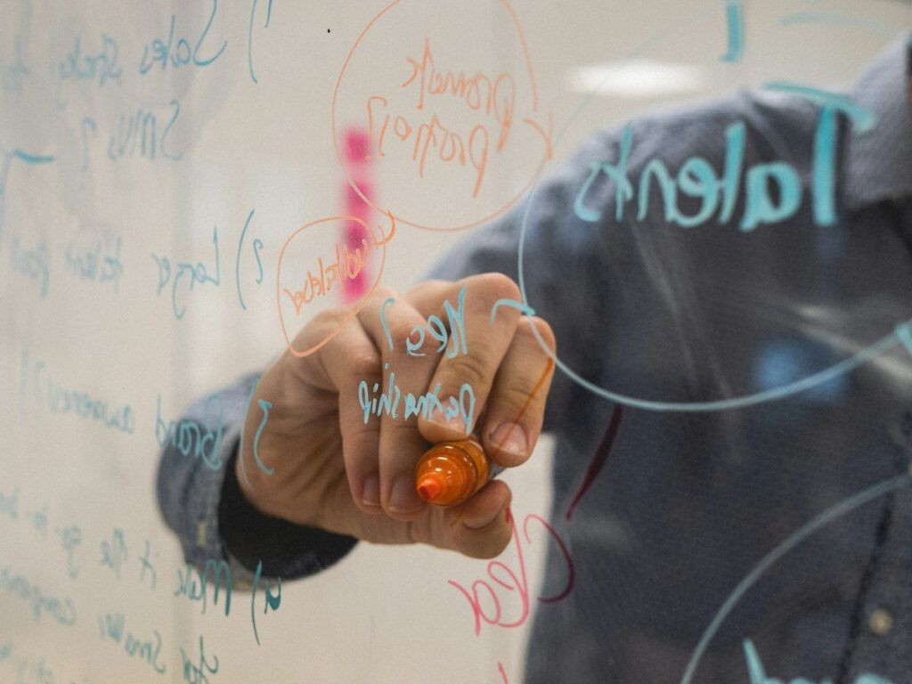 A hand with an orange marker sketching ideas and diagrams on a glass whiteboard