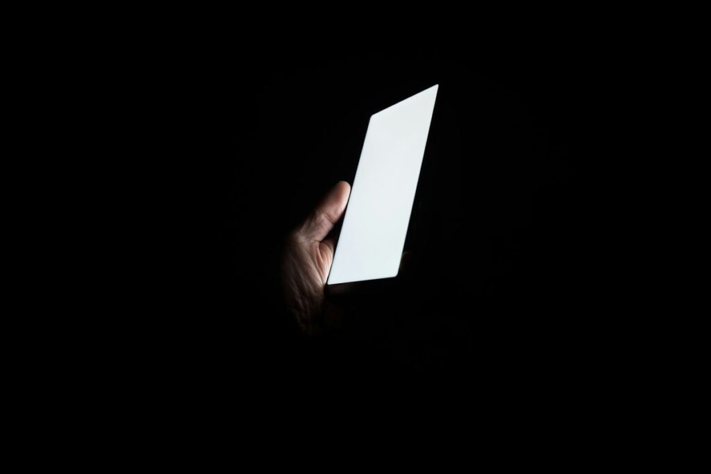 A hand holding a smartphone whose bright white screen glows in total darkness