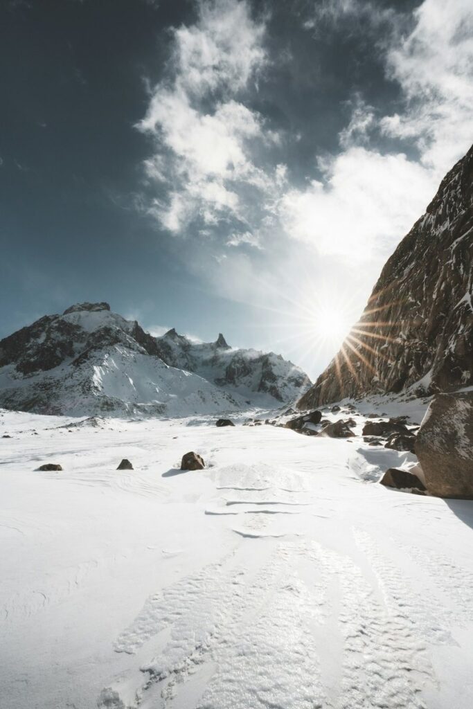 The snow-covered Mer de Glace glacier below jagged peaks of the Mont Blanc massif, with sun bursting past a dark rock wall under a brooding sky