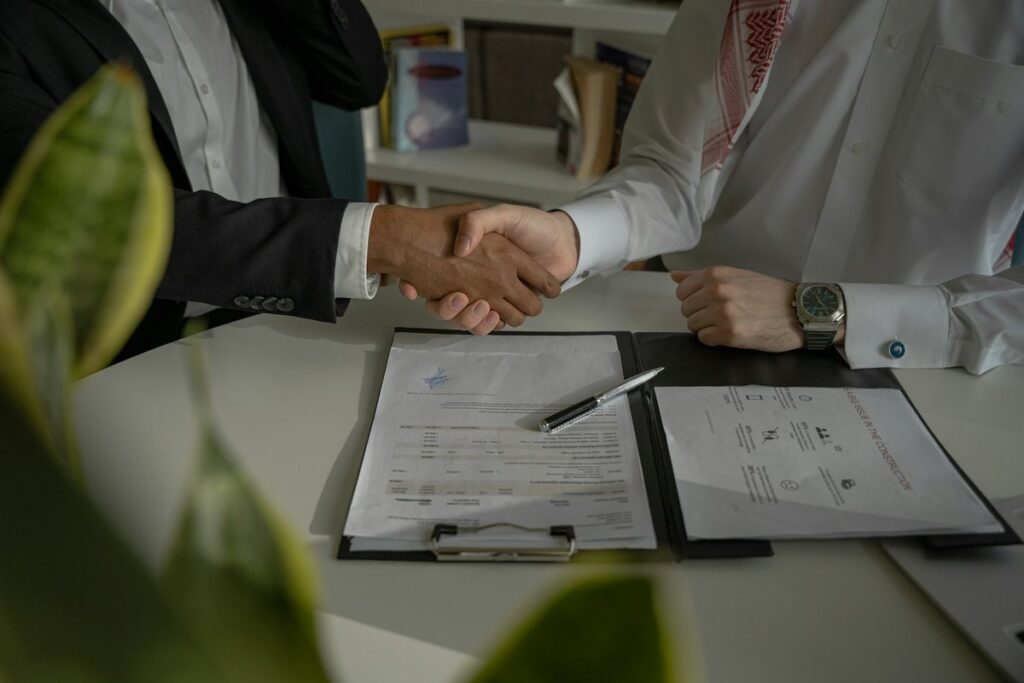 Two negotiators, faces out of frame, shaking hands across a table over a folder of contract papers and a pen