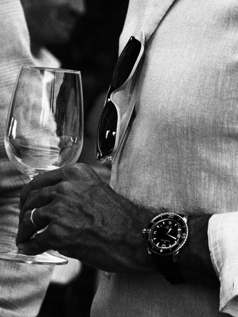 Black-and-white close-up of a man's hand holding a wine glass, with a luxury dive watch on his wrist and sunglasses tucked into his linen jacket