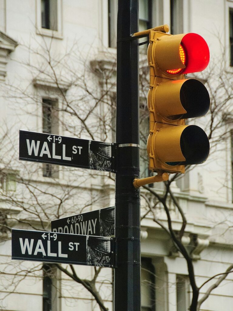 Wall St and Broadway street signs in Lower Manhattan beneath a traffic light glowing red
